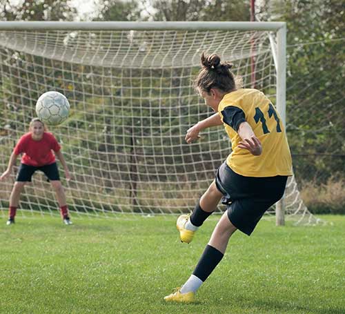 Two girls playing soccer. One girl kicks a ball into the net.