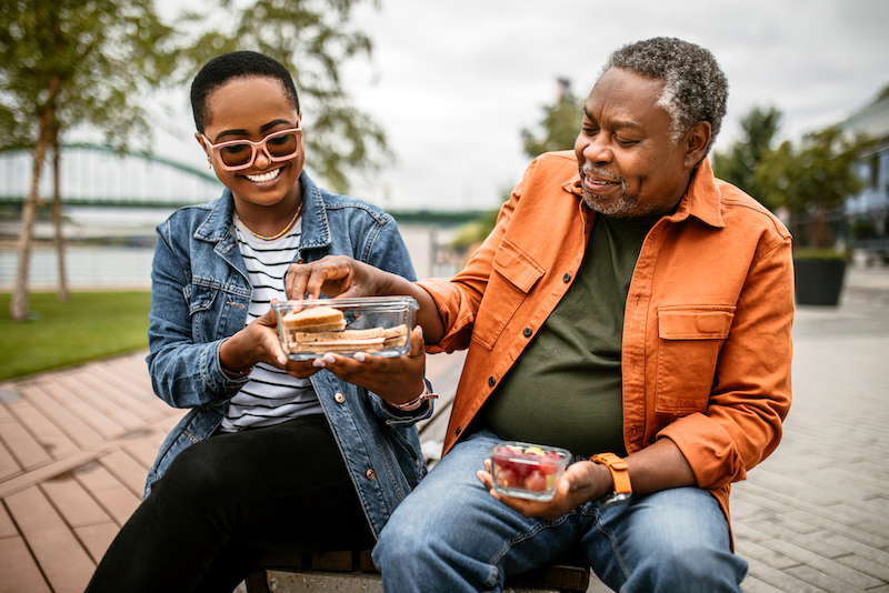 Father and adult daughter sitting outside in an urban park and sharing a picnic lunch.