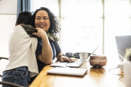 Mujer sentada en casa en una mesa trabajando en su computadora portátil y sonriendo mientras su hijo preadolescente la abraza.