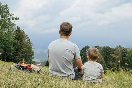 Rear view of a father and young son sitting in the grass on a cloudy day.