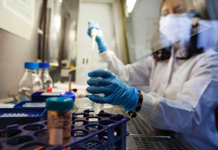 Close-up of a lab researcher's gloved hands, while they are holding a test tube and pipette inside a fume hood.