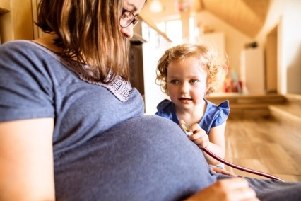 A toddler girl holding a stethoscope on her pregnant mother's belly.