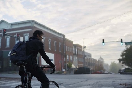 Un joven latino con mochila se sienta en su bicicleta frente a un semáforo verde, con una expresión seria en el rostro y las manos agarradas al manillar de la bicicleta.