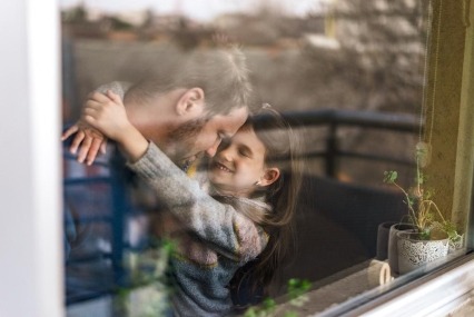View through a window of father and young daughter hugging and smiling at each other.