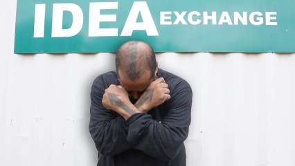 Screen shot of Saving Lives with Peer Support video: IDEA Exchange; photo of Chetwyn "Arrow" Archer with his head bowed down and arms crossed on his chest.  He has a down arrow drawn on his head and an up arrow on each hand and arm.