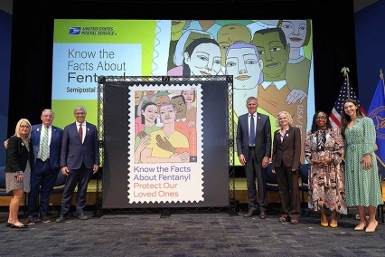 Seven participants stand onstage next to a large display of the U.S. Postal Service’s “Know the Facts About Fentanyl" semipostal stamp campaign. From left to right: Mary Ternan and Ed Ternan of Song for Charlie; Jay Bhattacharya of the NIH; Peter Pastre of the U.S. Postal Service; Nora Volkow of NIDA; Tamika Smith of WAMU; and mezzo-soprano Matilda Smolij. 