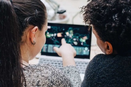  rear view of two adults sitting side-by-side at an office desk and talking over an open laptop computer.