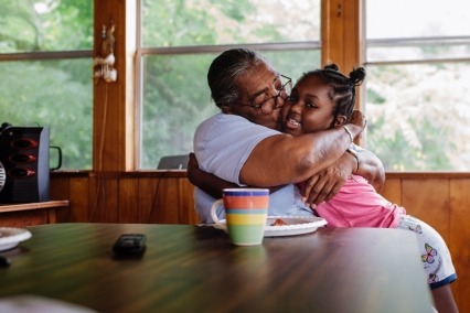 Grandmother sitting at the kitchen table and hugging young granddaughter.
