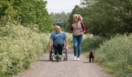 Un hombre con paraplejía en silla de ruedas y una mujer caminando, en un día soleado de verano en una reserva natural con su perro.