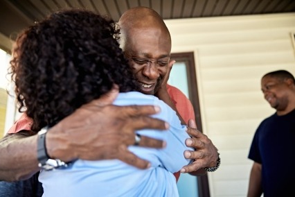 Older black man smiling and hugging a woman while standing outside on the front porch of a home with a group of people