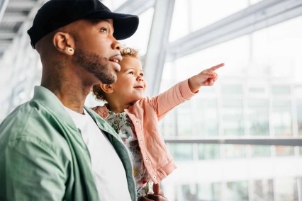 A child points excitedly out of an airport window onto the airfield while being held by an attentive father, both sharing a moment of discovery.