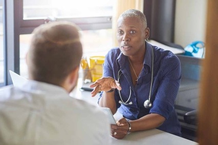 Una médica se sienta en su escritorio y conversa con un paciente masculino mientras mira los resultados de su prueba en su tableta digital.