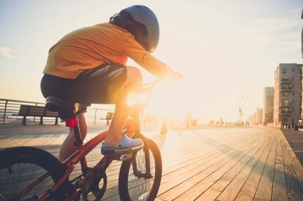 A young boy riding his bicycle on the boardwalk by the beach backlit by the sun wearing a helmet.