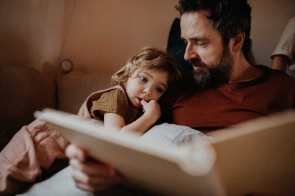 Father sitting with his young daughter resting on his arm at home, looking at a photo album.