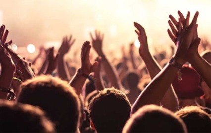 Audience with hands raised at a music festival and lights streaming down from above the stage.