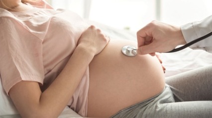 Pregnant woman resting on bed while doctor touches stethoscope to her abdomen.