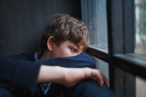 : Sad teenaged boy sitting at the window covering his face with his arm.