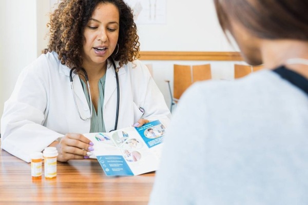 Médico sentado en su escritorio con un paciente compartiendo un folleto mientras habla sobre medicamentos recetados.