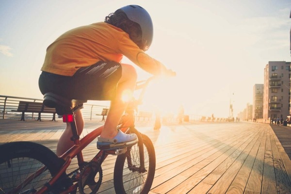 A young boy riding his bicycle on the boardwalk by the beach backlit by the sun wearing a helmet.