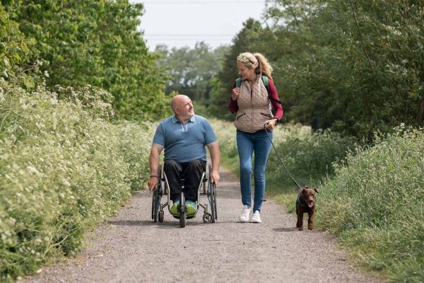 A man with paraplegia in a wheelchair and a woman walking, on a day out in a nature reserve with her dog on a sunny summer's day.