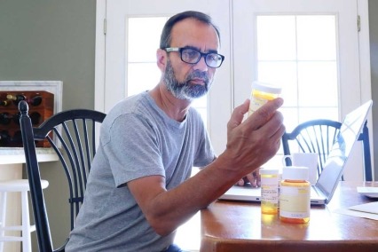 Man sitting at the dining room table looking at the label on a prescription medication bottle.